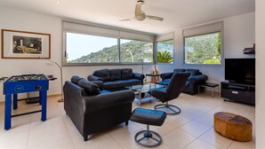 A modern living room with a foosball table, two sofas, a glass coffee table, a TV, and large windows with a view of greenery. - Exclusive Villa with Pool, Sea View, and Wi-Fi (Tossa De Mar)