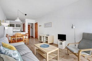 A living room with a sofa, a coffee table, a TV, a chair, a dining table with chairs, and a kitchen area in the background.