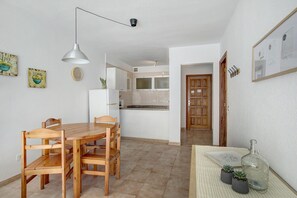 A dining area with a wooden table and chairs, a pendant light, a refrigerator, and a kitchen in the background.