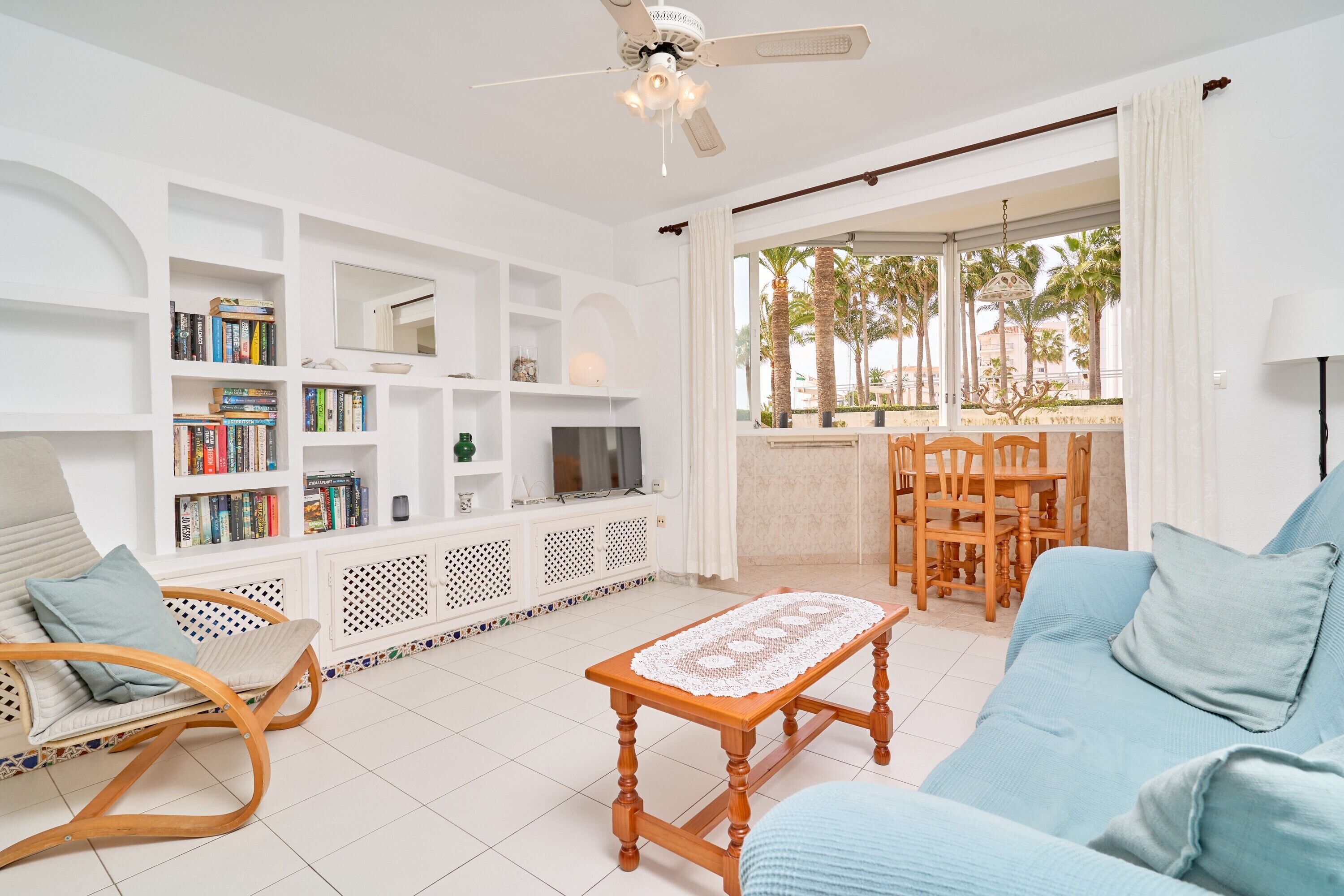 A living room with a sofa, a wooden coffee table, a bookshelf, a TV, and a dining area with chairs.