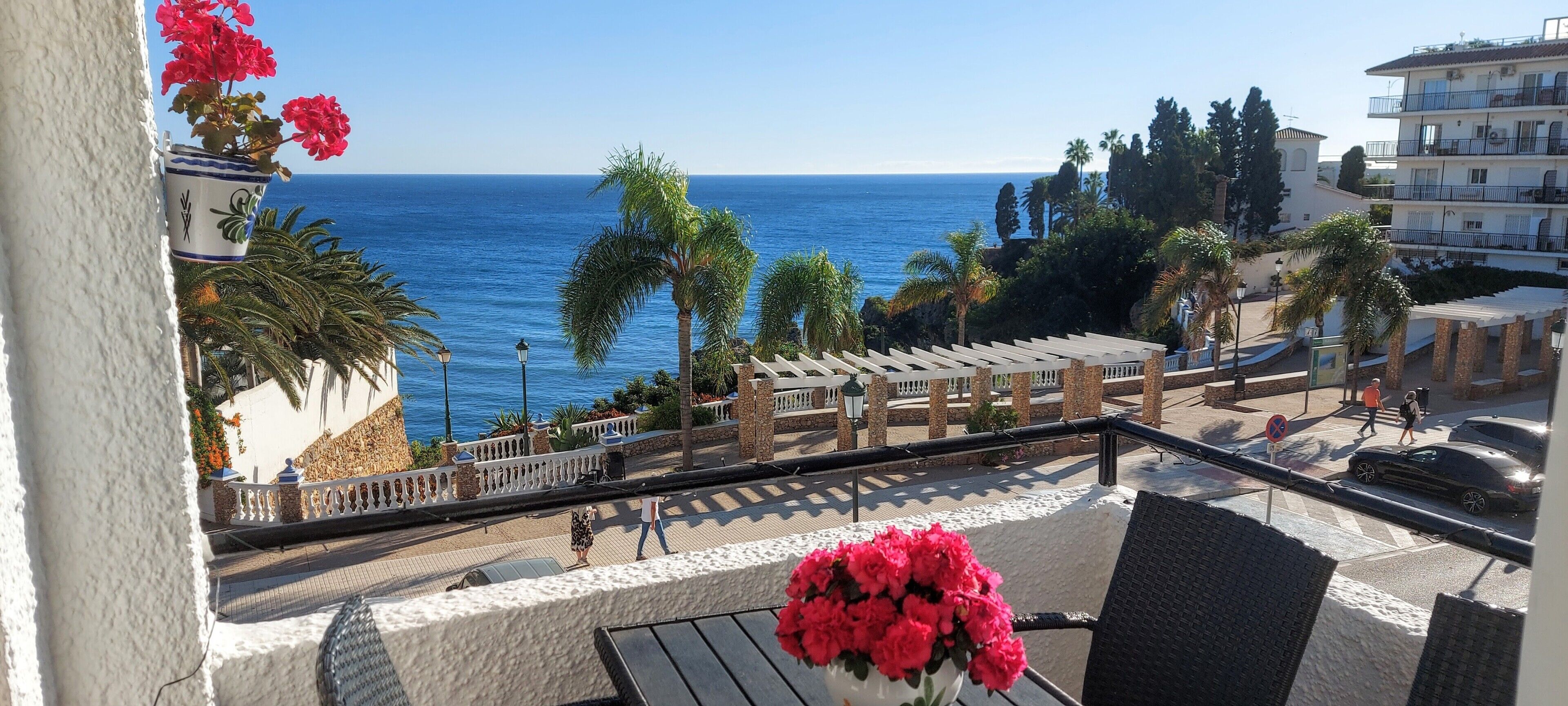 A balcony with a view of the sea, palm trees, and a white building with a balcony.