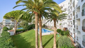 Un hotel de varios pisos con piscina rodeada de palmeras y un césped cuidado.