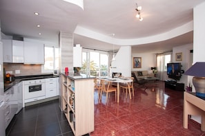A modern kitchen with white cabinets, a dining area with a round table and chairs, and a living room with a sofa and TV.