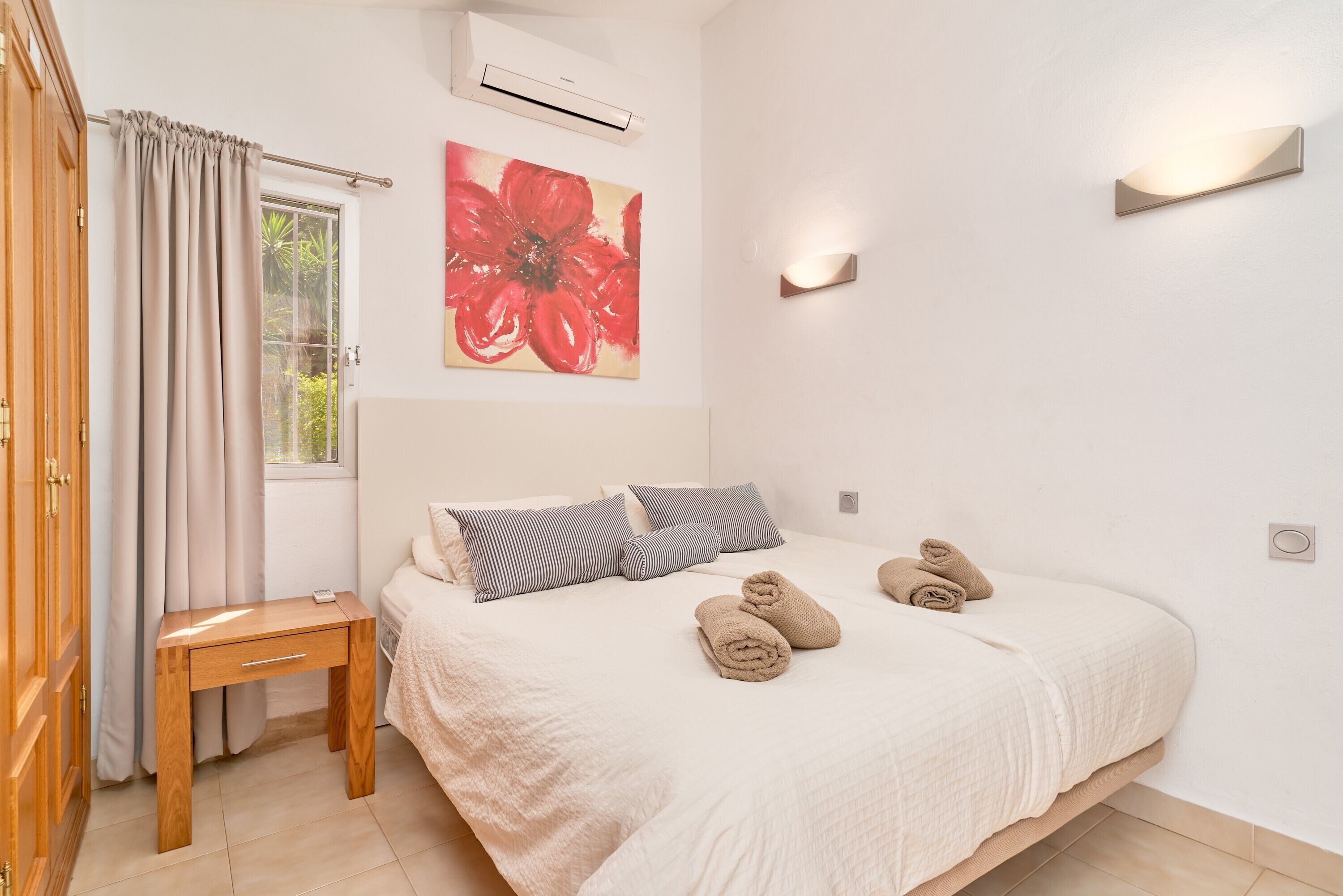 Een slaapkamer met een bed, kussens, een nachtkastje, een raam met gordijnen, een airconditioning en een wandlamp.