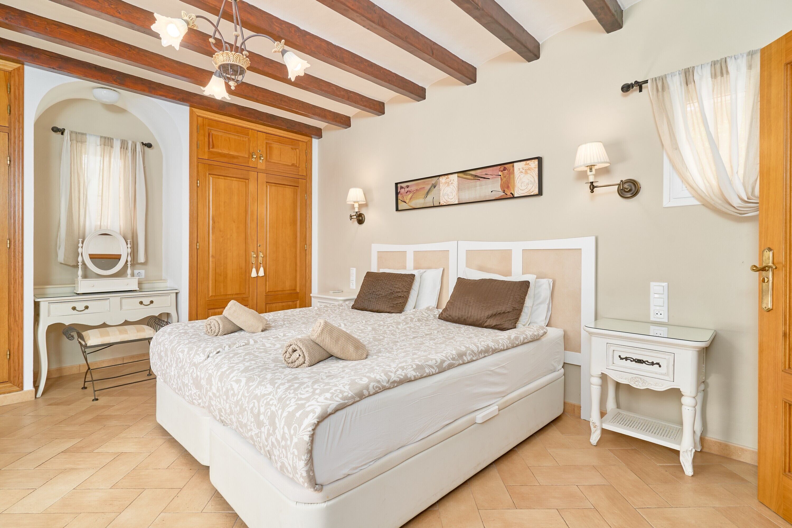A bedroom with a large bed, wooden wardrobe, bedside tables, and a ceiling with exposed beams.