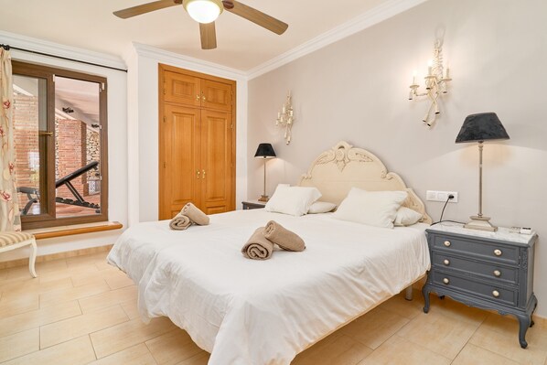Une chambre à coucher comprenant un lit, des tables de chevet, une armoire en bois, un ventilateur de plafond et une vue sur un mur de briques, visible à travers une porte vitrée.