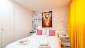 Quarto de hotel com cama, almofadas, mesa de cabeceira, um quadro e cortina.