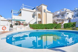 Une piscine à l’eau d’un bleu cristallin, entourée d’un bâtiment blanc doté de balcons et d’une haie verdoyante.