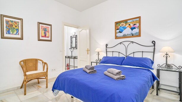 Un dormitorio con una cama, dos mesitas de noche, una silla y obras de arte en las paredes.