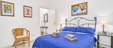 Un dormitorio con una cama, dos mesitas de noche, una silla y obras de arte en las paredes.