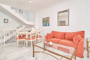 A living room with a staircase, a glass coffee table, a sofa, and two paintings on the wall.