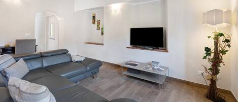 Un soggiorno moderno con un divano ad angolo grigio, una TV a schermo piatto montata a parete, un piccolo tavolino e una pianta decorativa.