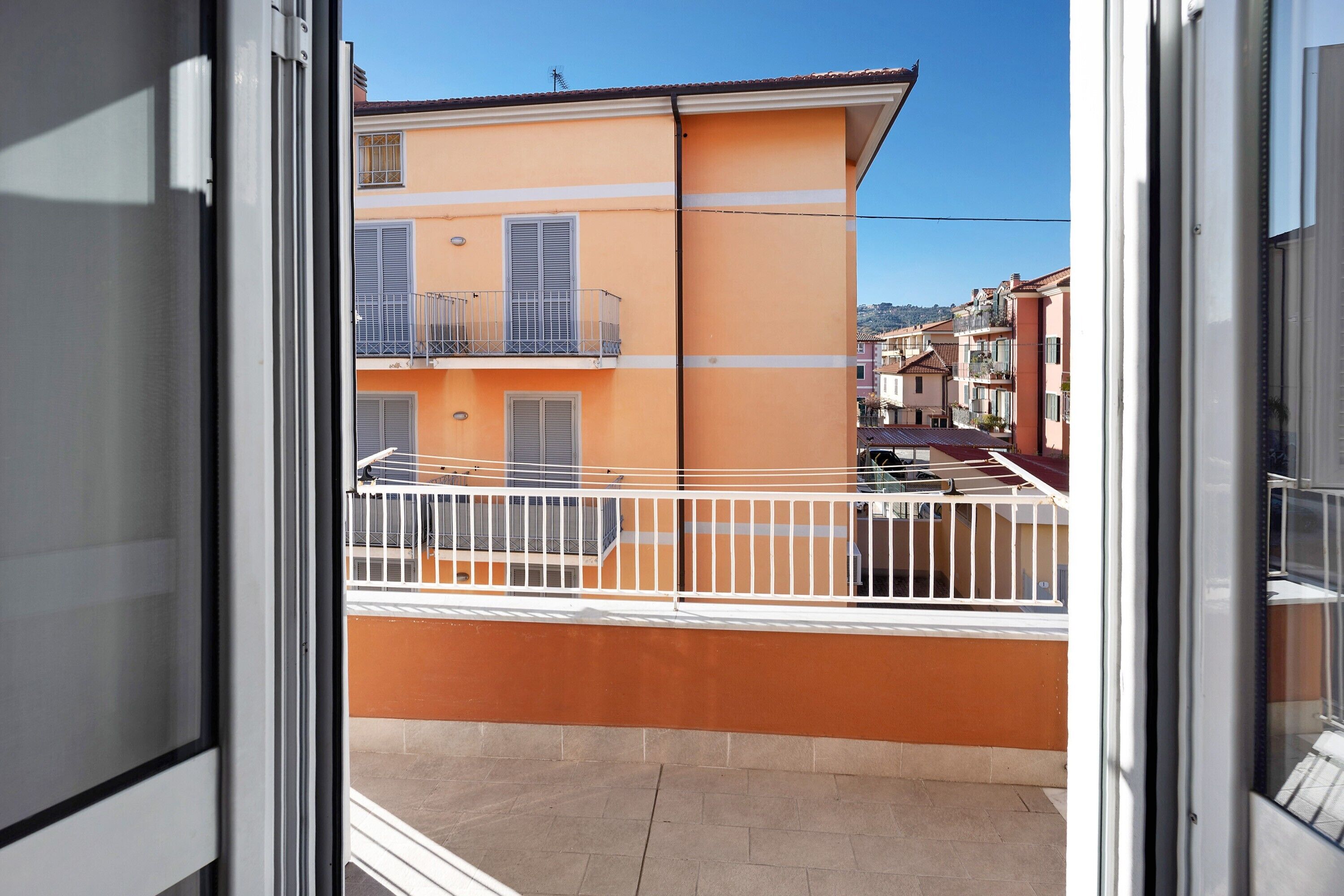 Un balcon avec une balustrade blanche, un immeuble aux murs orange et un ciel dégagé.