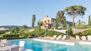 Piscina com espreguiçadeiras e mesa, rodeada por vegetação e um edifício de pedra ao fundo.
