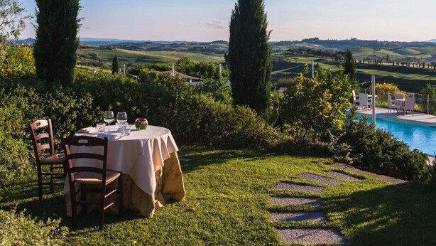 Mesa de jantar montada ao ar livre, com vista para uma piscina e colinas ondulantes ao fundo.