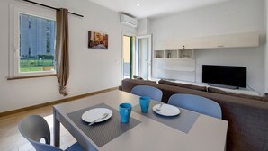 A modern room with a dining table set for two, a sofa, a flat-screen TV, and a window with curtains. - Ca di perti Apartment 1 with Wi-Fi, A/C & Garden (Finale Ligure)