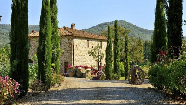 A stone building with a gravel driveway, surrounded by tall cypress trees and flowering plants. - Tuscan Family Villa with Sauna (Bucine)