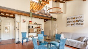 A dining area with a glass table and blue chairs, a sofa, a chandelier, and a decorative wall piece.