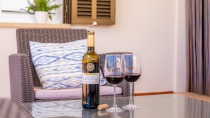 Une bouteille de vin et deux verres sur une table en verre, dans une pièce où se trouvent un canapé en osier et un coussin à motifs.