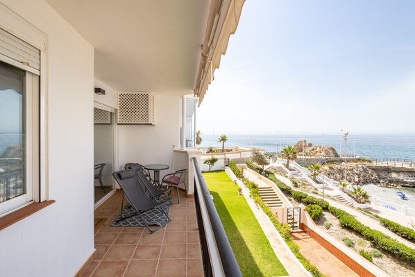 A balcony with a view of the sea, a small table with chairs, and a tiled floor.