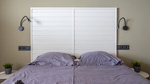 Quarto com cama grande, duas luminárias de cabeceira, uma tomada na parede e um pequeno vaso com planta.