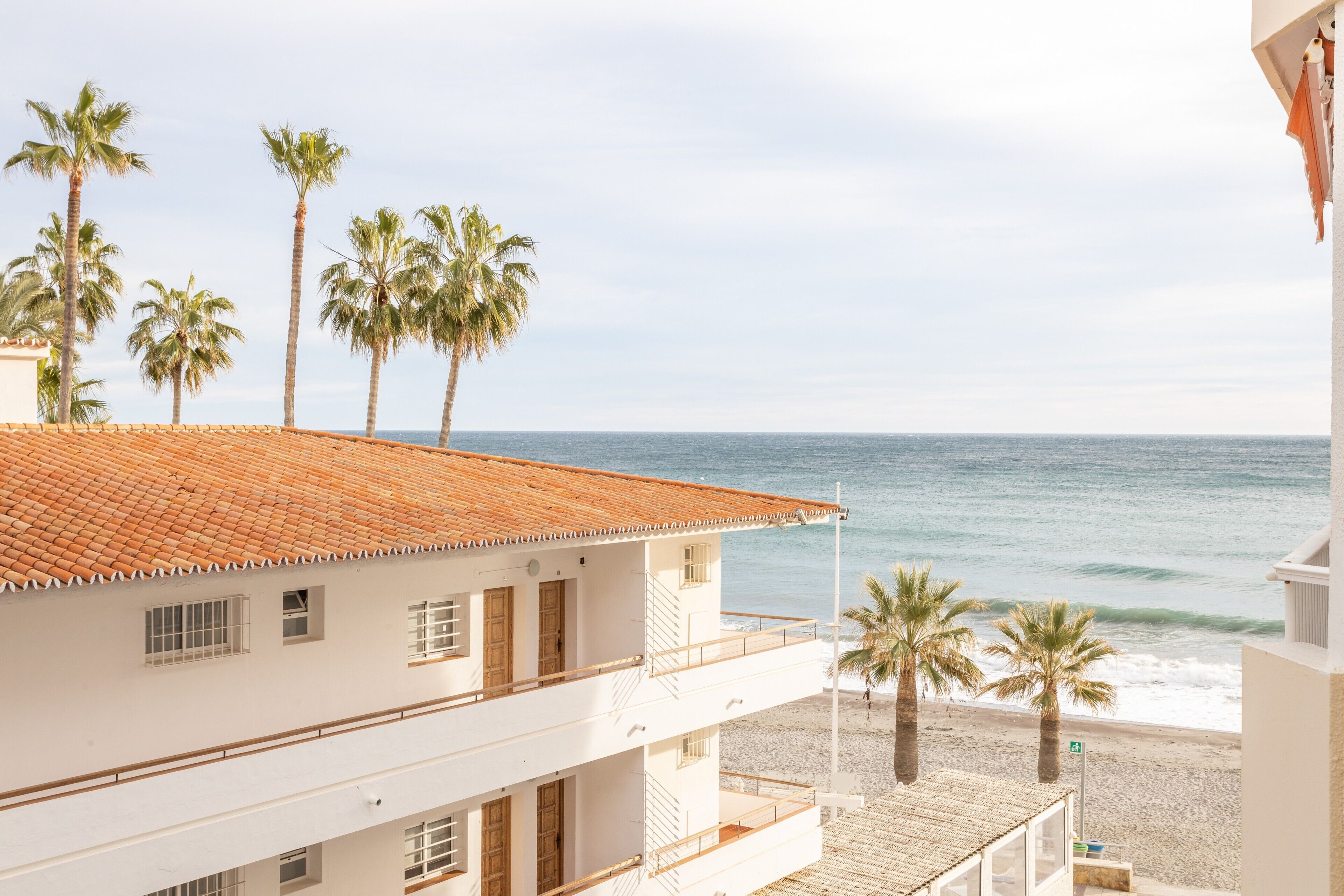 Ein Strandgrundstück mit einem Ziegeldach, zahlreichen Fenstern und einem Balkon mit Meerblick.