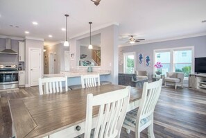 A modern kitchen with white cabinetry, a dining area with a wooden table and white chairs, a living room with a sofa and TV, and a balcony with a view.