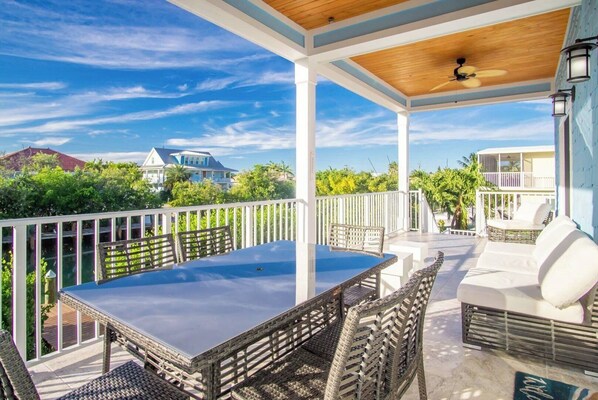A covered outdoor patio with a table and wicker chairs, white outdoor furniture, and a view of surrounding buildings and greenery.