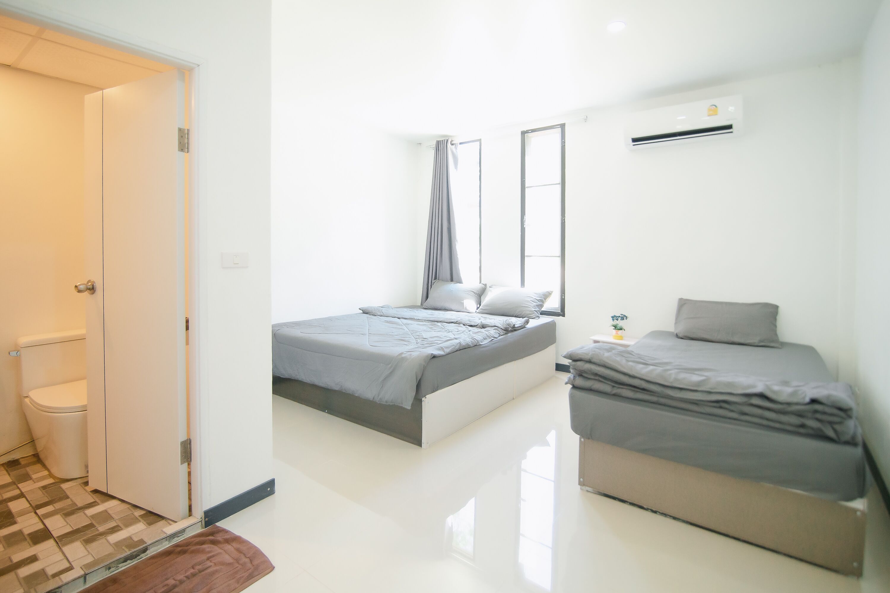A small, modern bedroom with a single bed, a second bed, a window with curtains, and an air conditioning unit.
