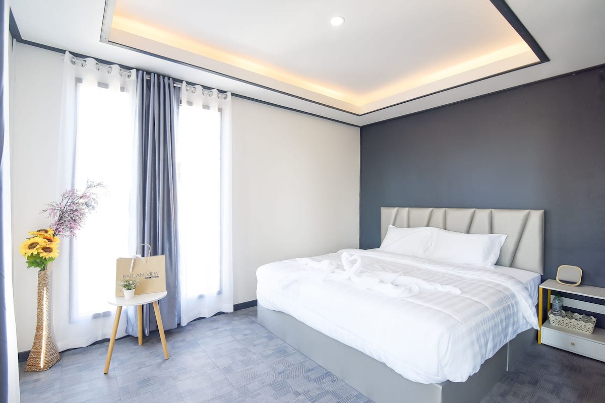 A modern hotel room with a large bed, a bedside table, a nightstand, a vase with flowers, and a view of the outdoors through the window.