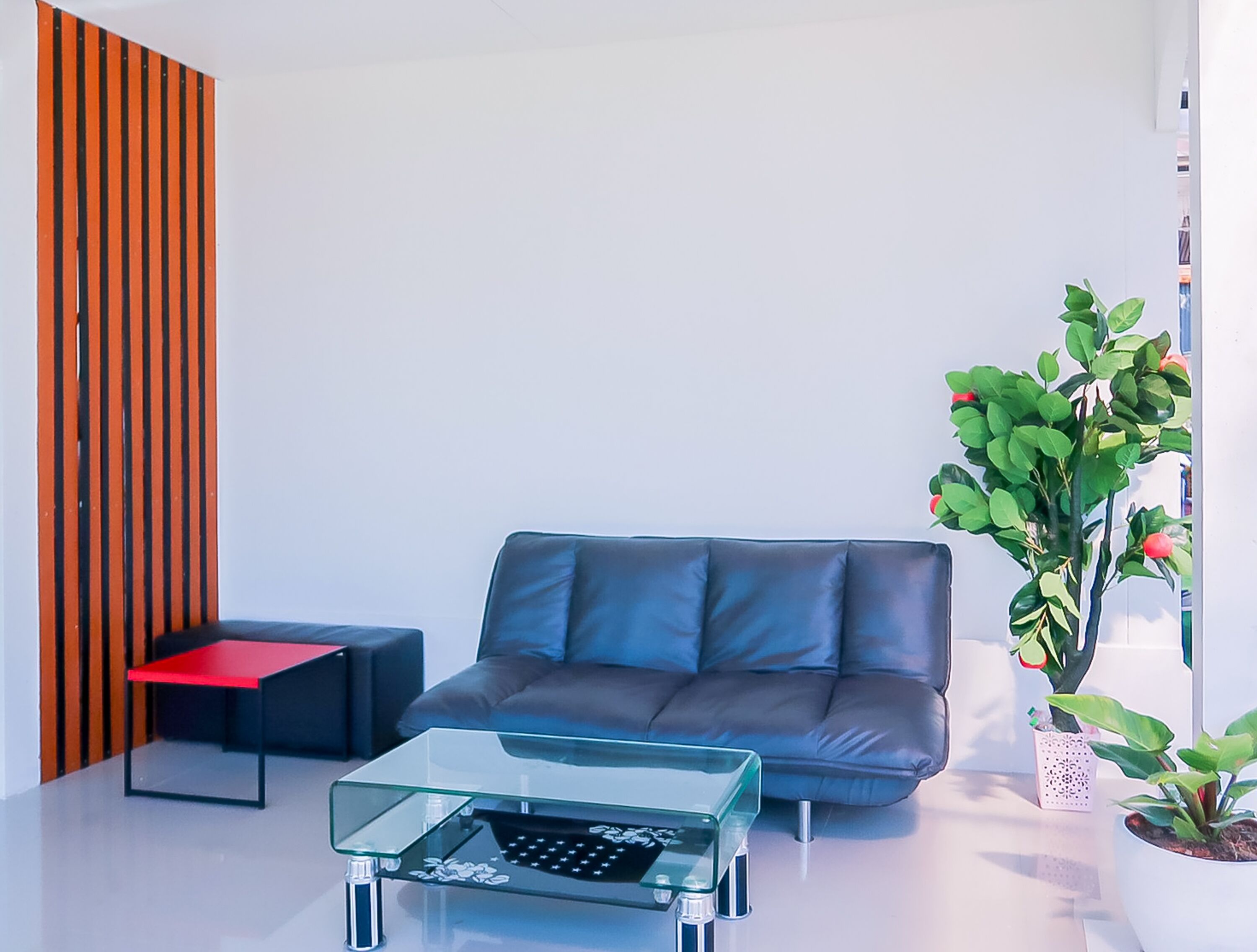 A modern living room with a blue sofa, a glass coffee table, a red side table, and a potted plant.