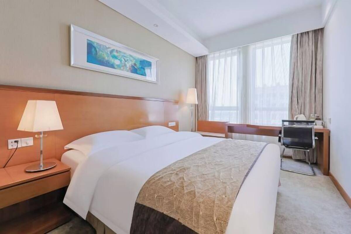 A hotel room with a bed, bedside lamps, a desk, a chair, a window with curtains, and a framed picture on the wall.