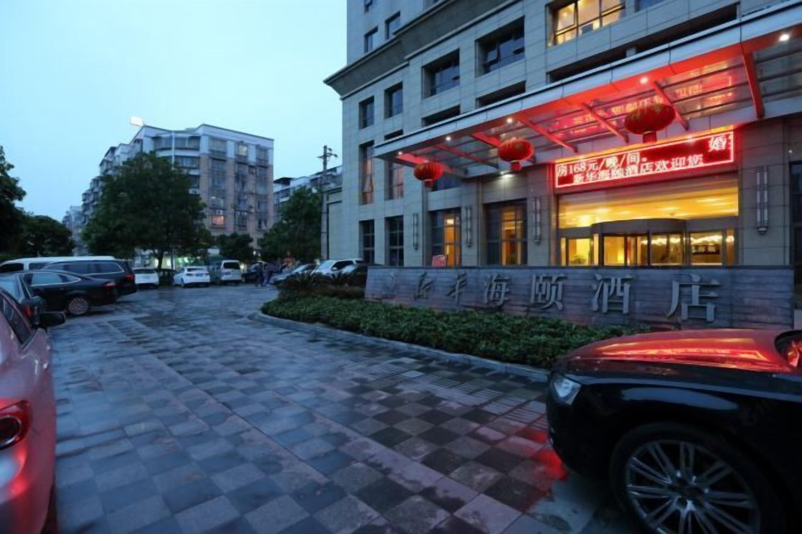 A hotel entrance with a checkered pavement, cars parked in front, and a building with illuminated signage.