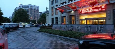 A hotel entrance with a checkered pavement, cars parked in front, and a building with illuminated signage.