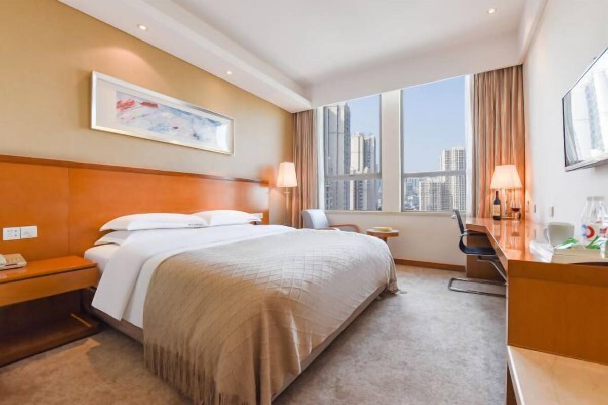 A hotel room with a large bed, a desk, a chair, a window with a city view, and a framed picture on the wall.