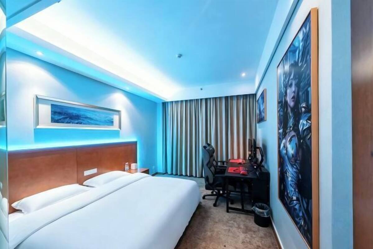 A hotel room with a bed, a desk with a chair, a painting on the wall, and a window with curtains.