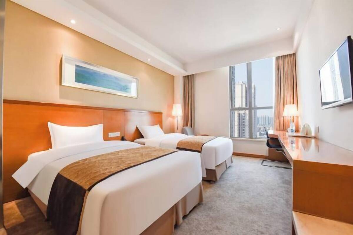 A hotel room with two beds, a desk, a chair, and a large window with a city view.