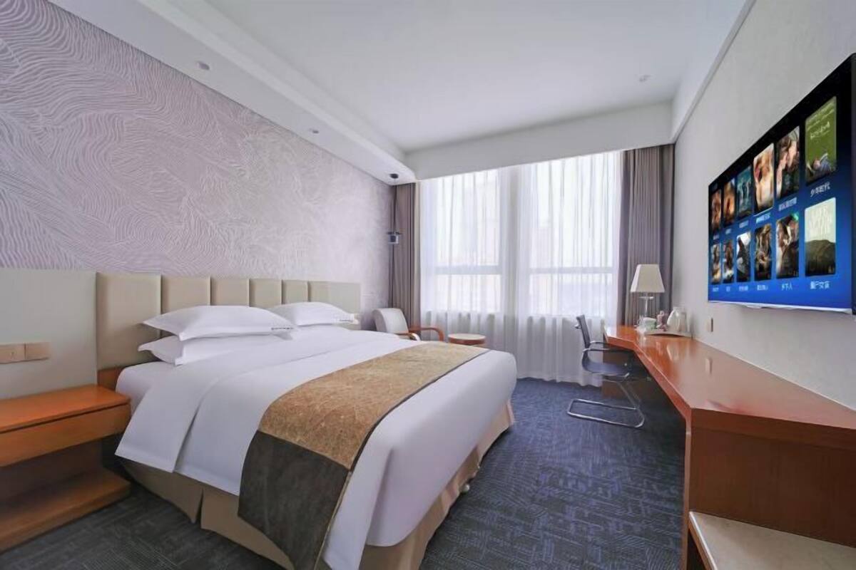 A hotel room with a large bed, a desk, a chair, a TV on the wall, and a window with curtains.