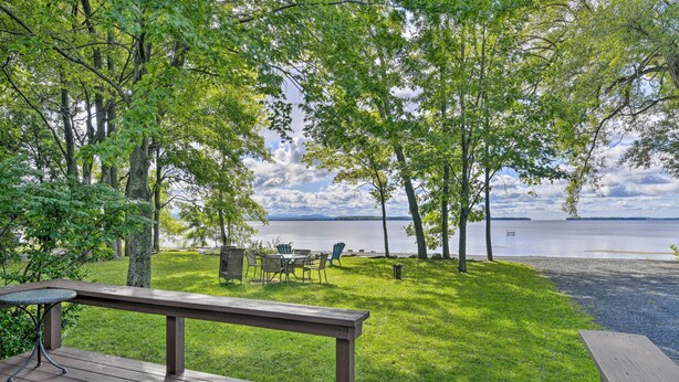 Een terras met tafel en stoelen, uitkijkend op een gazon met bomen en een meer.