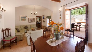 Un comedor con una mesa de madera, sillas y un jarrón con flores. Se observa un sofá y una mesa de comedor al fondo.
