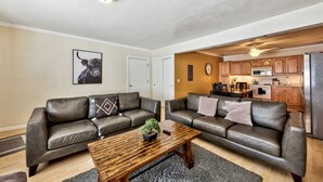 Una sala de estar con dos sofás de cuero, una mesa de centro de madera y una pequeña planta. Se ve una cocina al fondo, con armarios de madera y un refrigerador.