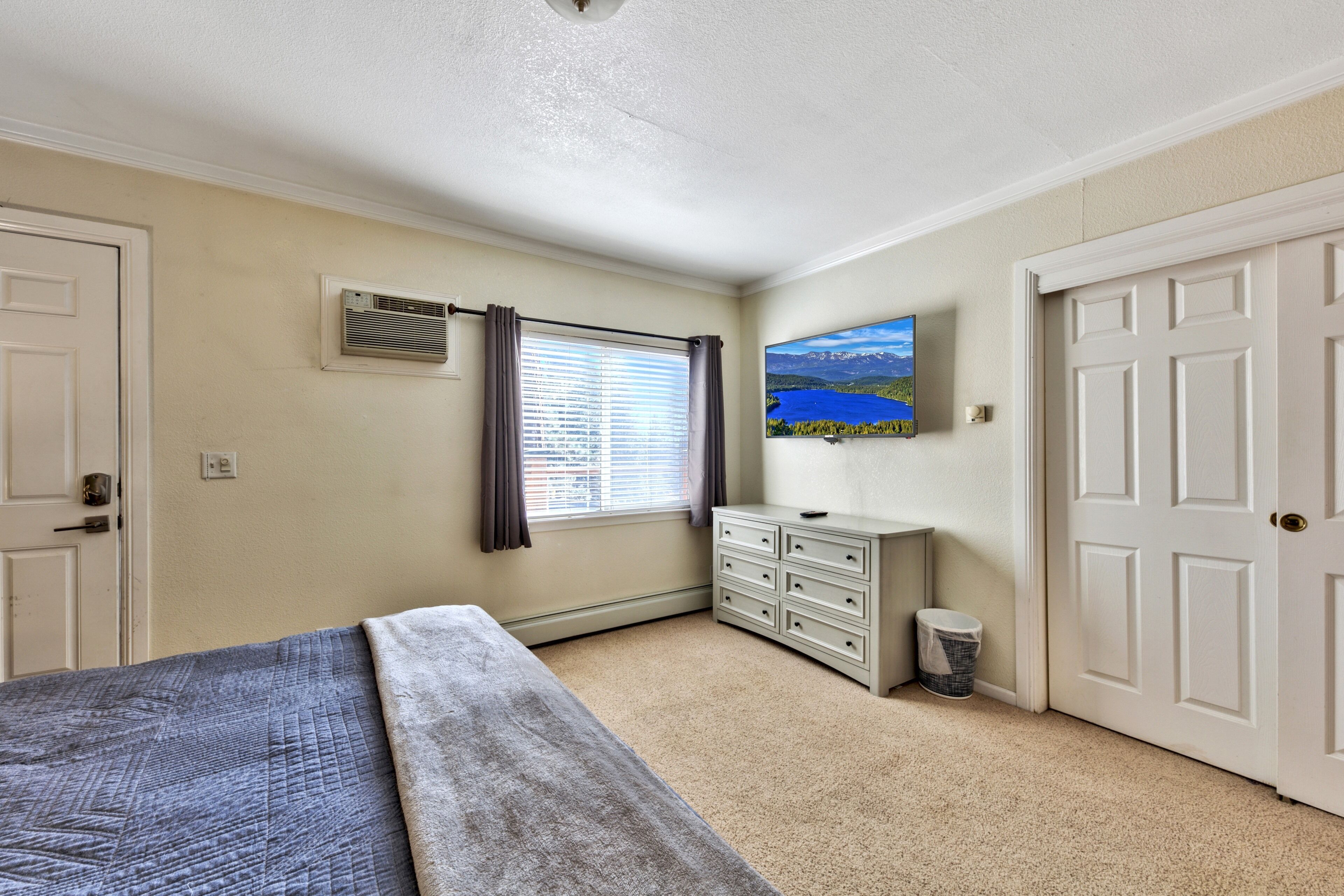 A bedroom with a bed, a dresser, a window with curtains, a wall-mounted TV, and a door.