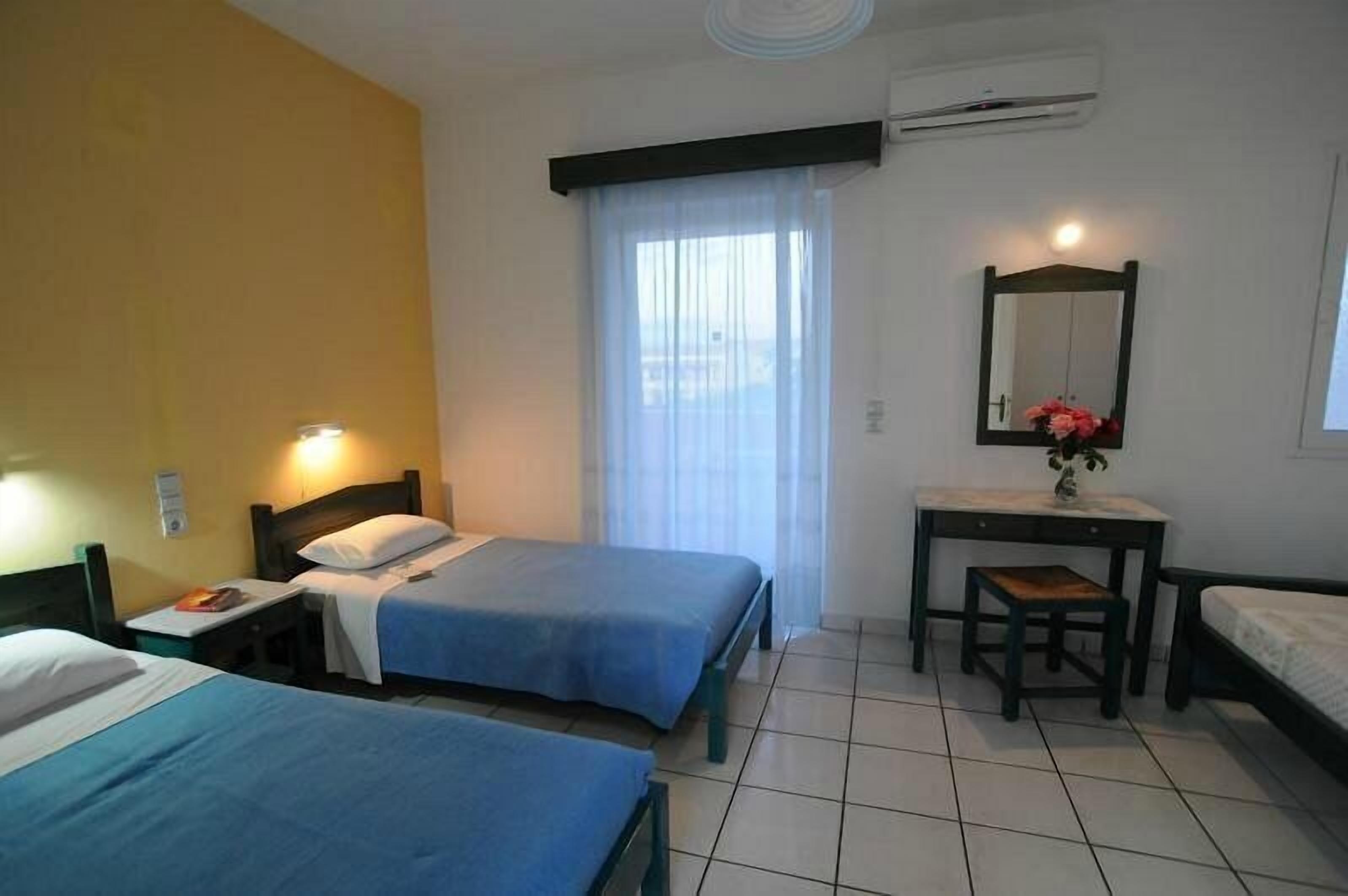 Een hotelkamer met twee bedden, een klein bureau, een spiegel en een raam met dunne gordijnen.