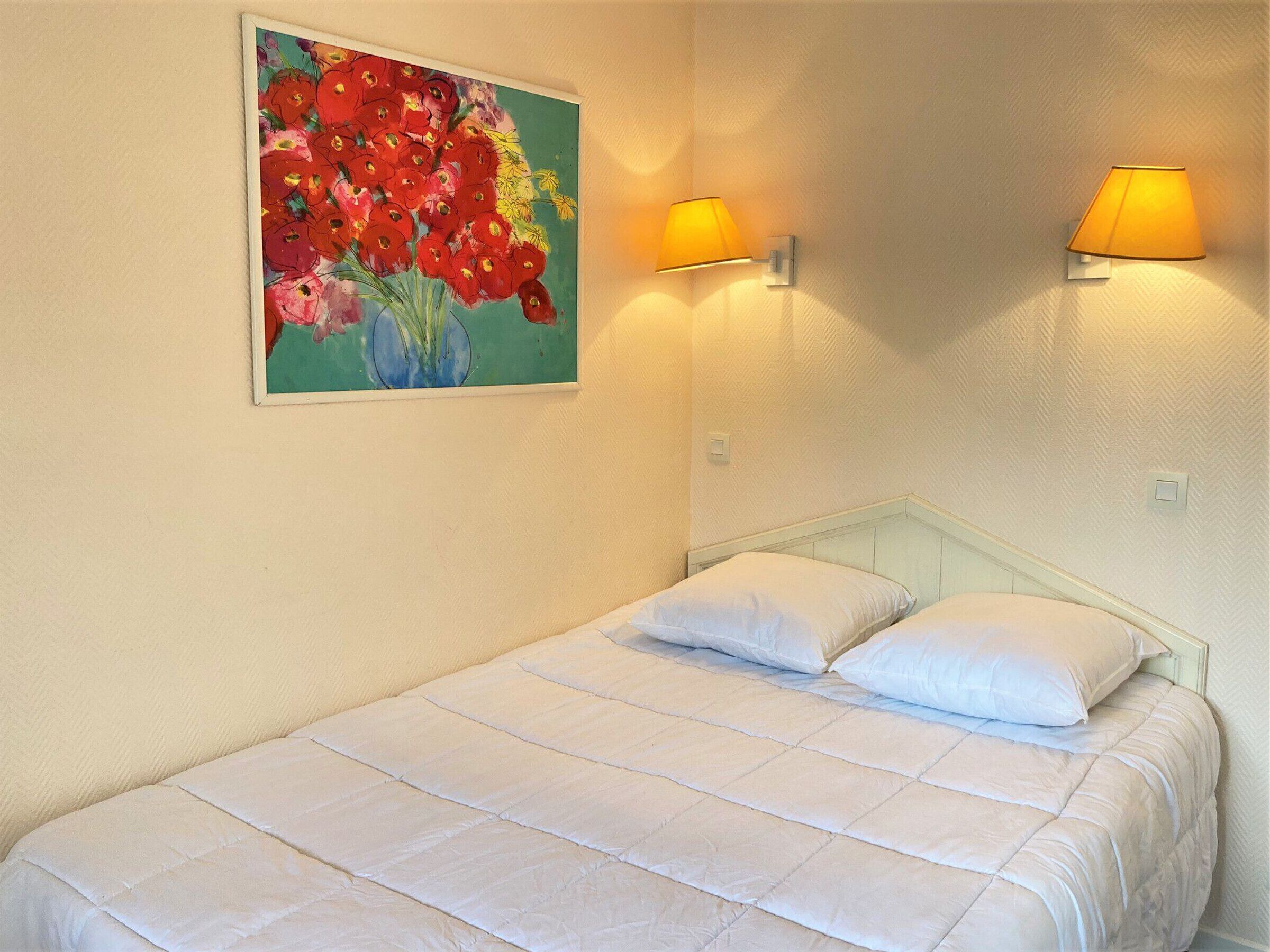 Un lit simple avec une literie blanche, deux lampes fixées au mur et un tableau représentant des fleurs accroché au mur.