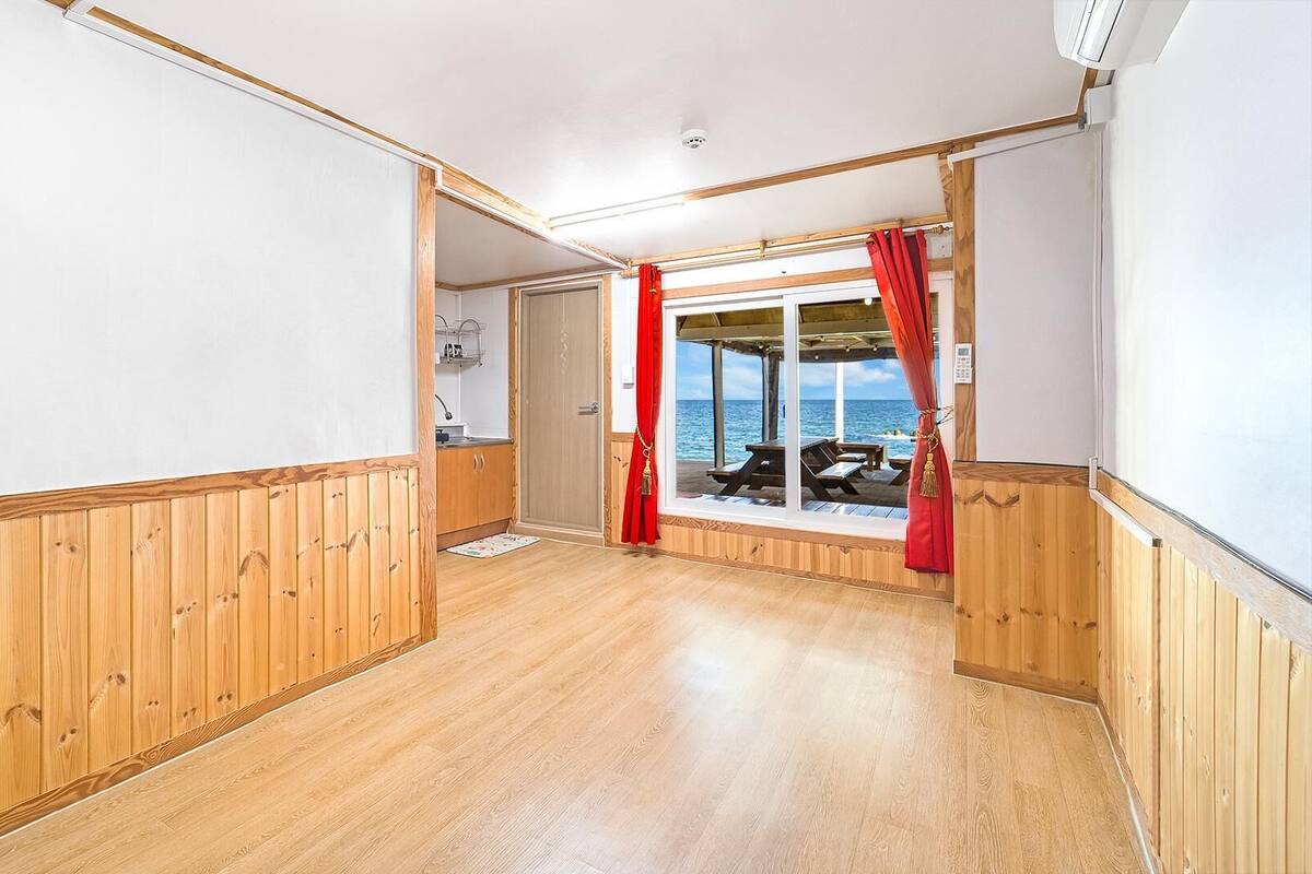 Ein Zimmer mit Holzwänden und Holzboden, einem Fenster mit roten Vorhängen und Blick aufs Meer.