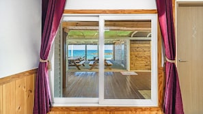 Une pièce dotée d’un plancher en bois, avec une porte-fenêtre coulissante donnant sur un espace extérieur aménagé avec des tables de pique-nique et offrant une vue sur la mer.