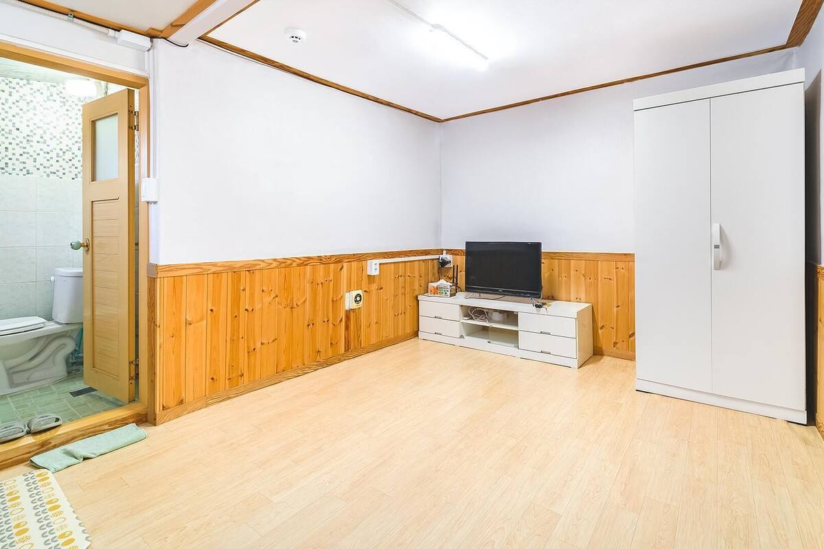 Ein kleiner, sauberer Raum mit Holzwänden, einem weißen Kleiderschrank, einem Fernseher auf einem weißen Schrank und einer durch eine Glastür sichtbaren Toilette.