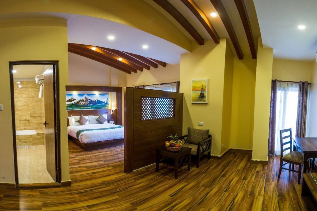 Une chambre d’hôtel avec un plancher en bois, un lit orné d’un tableau représentant une montagne, un coin repas avec une table et des chaises, et un petit coin salon avec un panier de fruits.