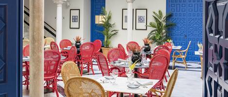 A dining area with red and wicker chairs, white tables, and a blue door.