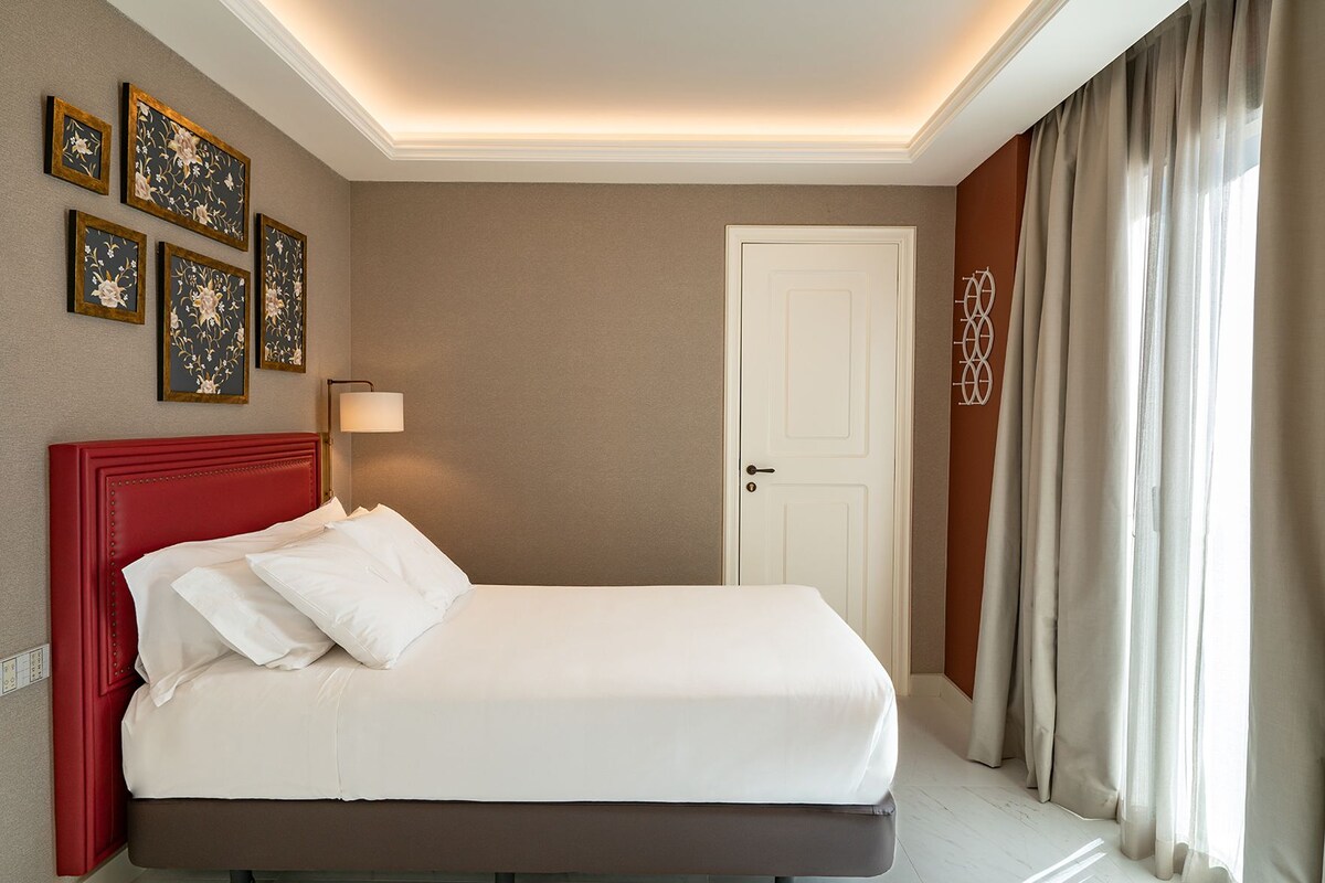A hotel room with a red headboard, white bedding, a door with a decorative handle, and a wall with framed artwork.